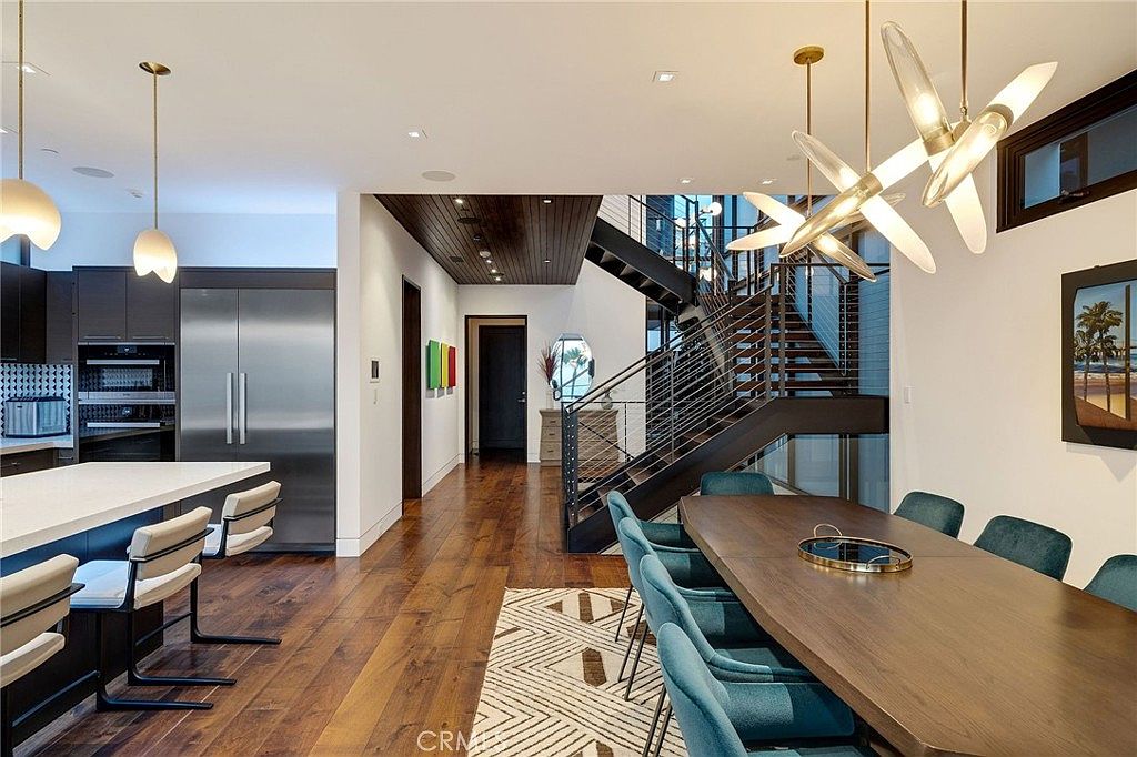 This open-concept space seamlessly blends a modern kitchen with a formal dining area, featuring rich dark wood flooring throughout. The kitchen is equipped with stainless steel appliances and a large island, while the dining area is anchored by a long table and a striking, multi-armed contemporary chandelier. A dramatic staircase with metal railings leads to the upper level, creating a sophisticated and airy architectural focal point.