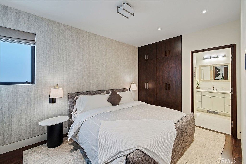 This contemporary bedroom features a neutral color palette with textured wallpaper and dark wood cabinetry. A plush upholstered bed sits centered in the room, flanked by minimalist bedside tables and wall-mounted lamps. The space flows directly into an adjacent bathroom, creating a cohesive and functional layout.
