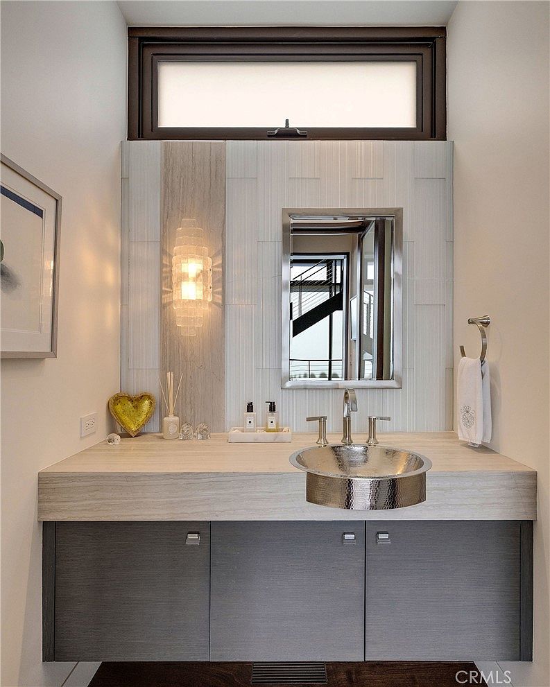 This contemporary powder room features a floating vanity with a dark wood-toned cabinet and a light-toned stone countertop. A unique hammered metal vessel sink sits atop the counter, complemented by a wall-mounted mirror and a decorative glass wall sconce. The space is accented by a textured tile backsplash and a high, horizontal transom window that allows natural light to filter into the room.