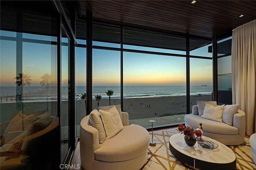 This sophisticated living space features floor-to-ceiling windows that frame a panoramic view of the ocean and beach at sunset. The room is furnished with curved, light-toned seating and a central oval coffee table, all set atop a patterned area rug. The dark wood-paneled ceiling adds a sense of warmth and luxury to the modern, open-concept design.