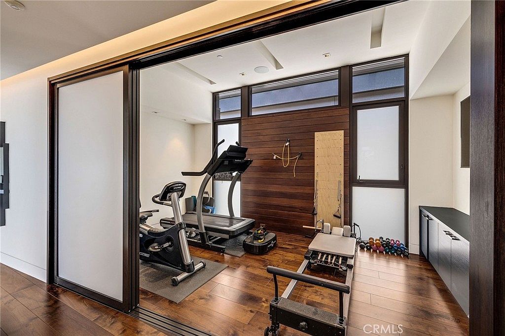 This modern home gym features dark wood flooring and a prominent wood-paneled accent wall, creating a sophisticated and focused workout environment. The space is equipped with a treadmill, a stationary bike, and a Pilates reformer, all positioned to take advantage of the natural light from the large windows. Sliding glass doors provide a seamless transition from the adjacent living area, emphasizing an open and airy architectural design.