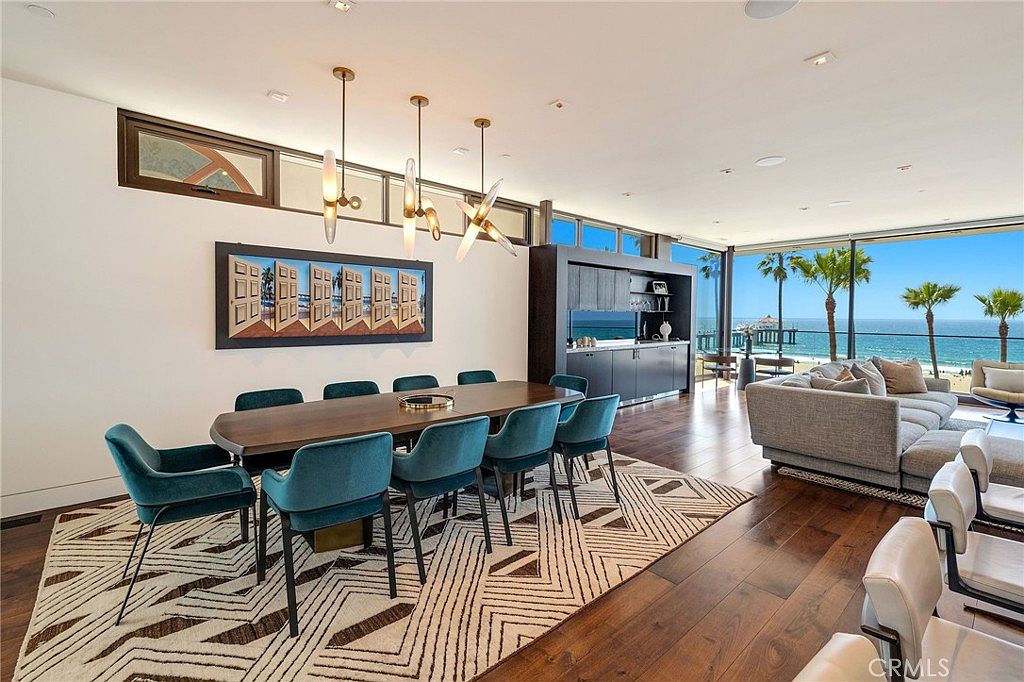 This open-concept dining area features a long, dark wood table surrounded by eight teal upholstered chairs, set atop a large geometric-patterned area rug. The space is illuminated by a modern multi-pendant light fixture and flows seamlessly into a living area with floor-to-ceiling glass walls that offer expansive ocean views. The room is finished with dark hardwood floors and a sleek, dark-toned built-in bar area, creating a sophisticated and airy coastal atmosphere.