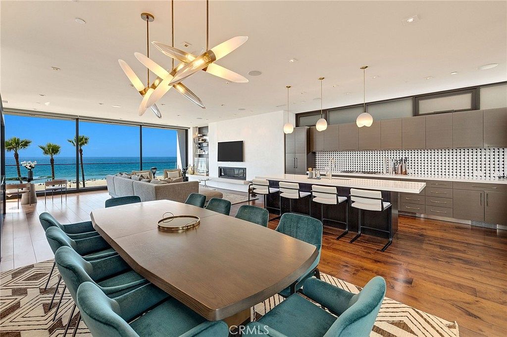 This open-concept living space features a large dining area in the foreground with a wooden table and teal upholstered chairs, transitioning into a spacious living room and kitchen. The room is defined by expansive glass walls that provide a panoramic view of the ocean and beach. A modern kitchen with a long island and bar seating sits to the right, while a minimalist fireplace is integrated into the wall near the living area.