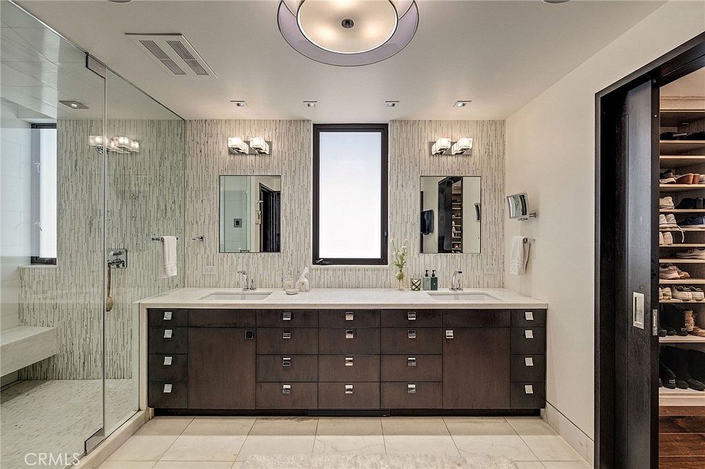 This primary bathroom features a dual-sink vanity with dark cabinetry and a light-toned countertop, set against a textured, vertical-patterned wall. A glass-enclosed shower is positioned to the left, while a sliding door on the right reveals a walk-in closet with organized shelving. The space is illuminated by a central dome light and wall-mounted fixtures above the mirrors, creating a clean and contemporary aesthetic.