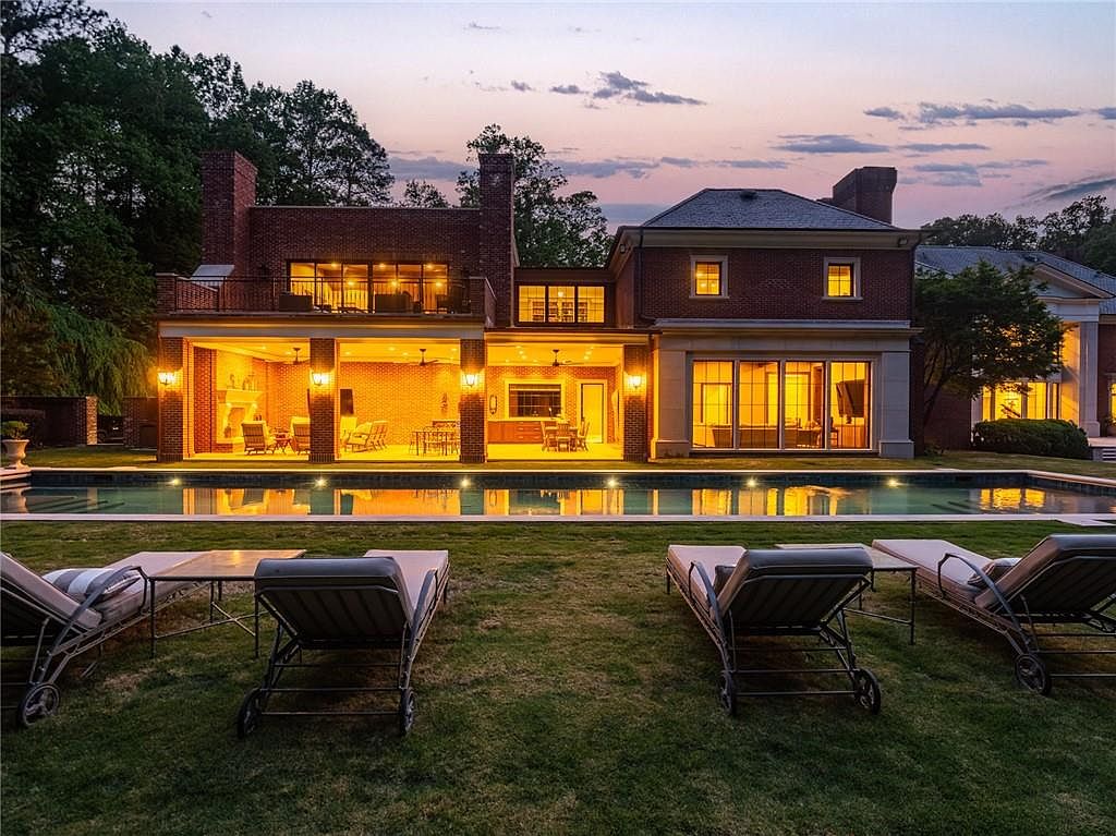 This elegant brick estate is captured at dusk, showcasing a sprawling rear facade with a large, illuminated covered patio and a rectangular swimming pool. The home features a mix of traditional brick architecture, expansive glass walls, and a second-story balcony, creating a sophisticated indoor-outdoor living experience. In the foreground, four lounge chairs are arranged on a manicured lawn, providing a serene vantage point of the glowing residence.