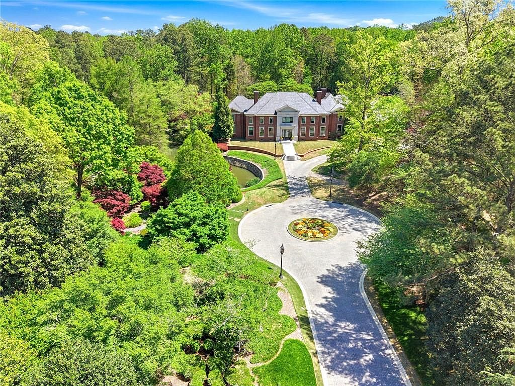 This high-angle aerial view captures a grand, multi-story brick estate nestled within a lush, mature forest. A sweeping, paved circular driveway with a central floral garden leads directly to the home's formal entrance, while a small, curved pond sits to the left of the structure. The property is surrounded by dense greenery, emphasizing a sense of privacy and seclusion in a park-like setting.