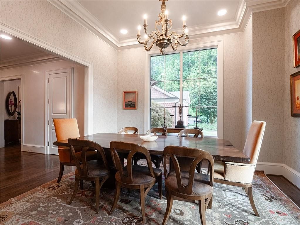 This elegant dining room features a large wooden table surrounded by a mix of upholstered and wooden chairs, set atop a detailed patterned area rug. A classic chandelier hangs above the table, while a large window provides a view of the exterior greenery and a decorative scale sculpture. The room is finished with textured wallpaper and crown molding, creating a sophisticated and formal atmosphere.