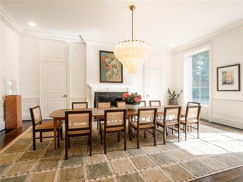 This elegant dining room features a long wooden table surrounded by eight chairs with woven cane backs and seats, set atop a large, multi-toned grid-patterned area rug. A striking, tiered feather-style chandelier hangs above the table, while a dark stone fireplace surround with a mantel serves as a focal point on the back wall. The room is brightened by natural light from large windows and is decorated with framed artwork, creating a sophisticated and inviting atmosphere.
