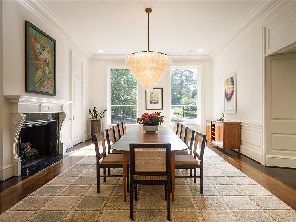 This elegant dining room features a long wooden table surrounded by cane-back chairs, centered beneath a striking tiered feather-style chandelier. To the left, a fireplace with a dark stone surround and a decorative mantel serves as a focal point, while a wooden sideboard sits against the right wall. Large windows at the far end of the room provide ample natural light and views of the lush greenery outside, all set upon a large, neutral-toned patterned rug.