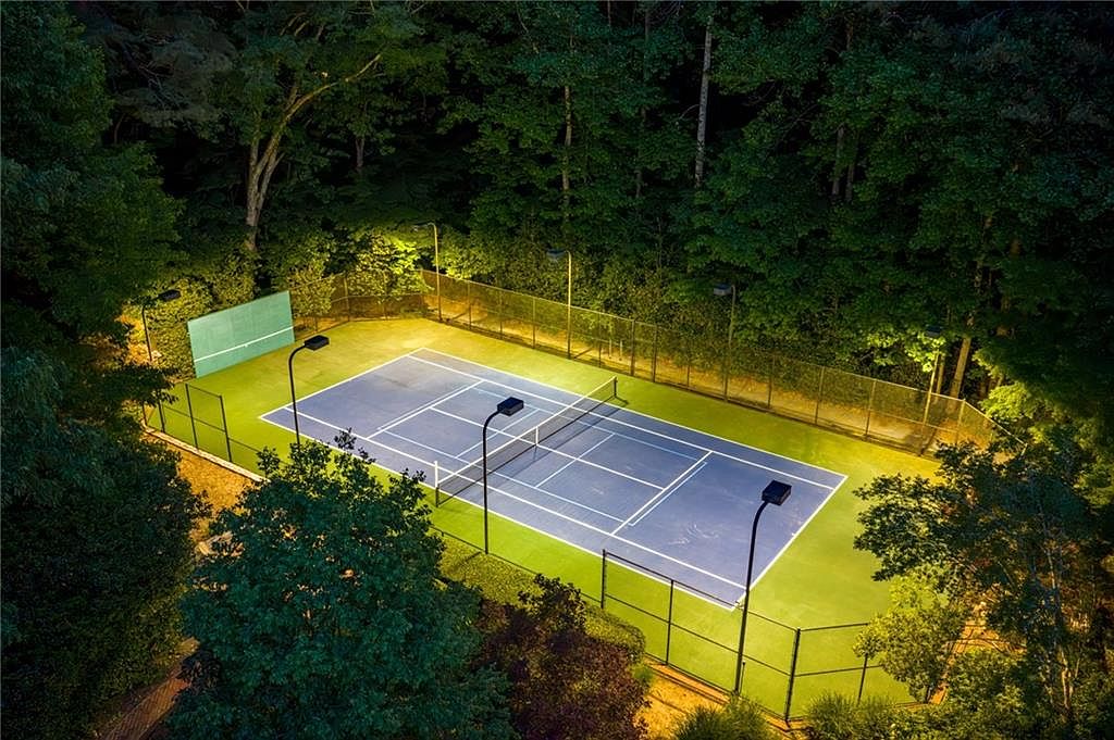 This high-angle aerial view captures a private, illuminated tennis court nestled within a dense, lush forest setting at night. The court features a blue playing surface with a practice backboard at one end and is fully enclosed by a tall chain-link fence. The scene is brightly lit by several tall stadium-style light poles, creating a dramatic contrast against the surrounding dark, wooded landscape.