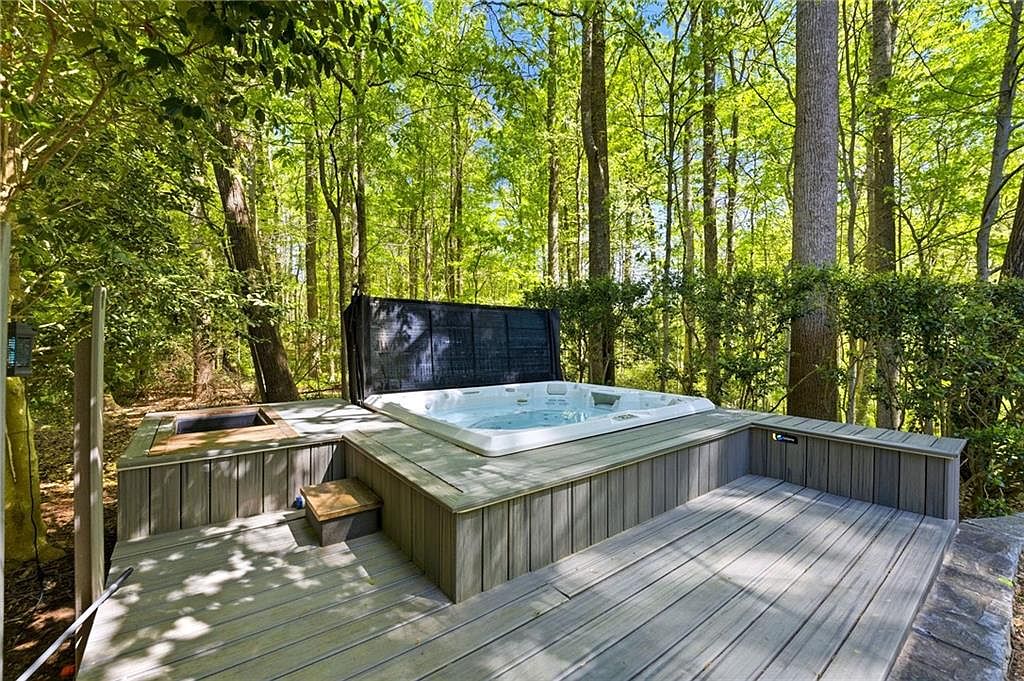 This outdoor space features a built-in hot tub set into a multi-level gray composite deck, surrounded by a lush, wooded forest. The deck includes a small integrated step and a recessed area, creating a private and serene retreat. The perspective is a wide-angle shot from the deck level, emphasizing the seamless integration of the spa into the natural, tree-filled landscape.