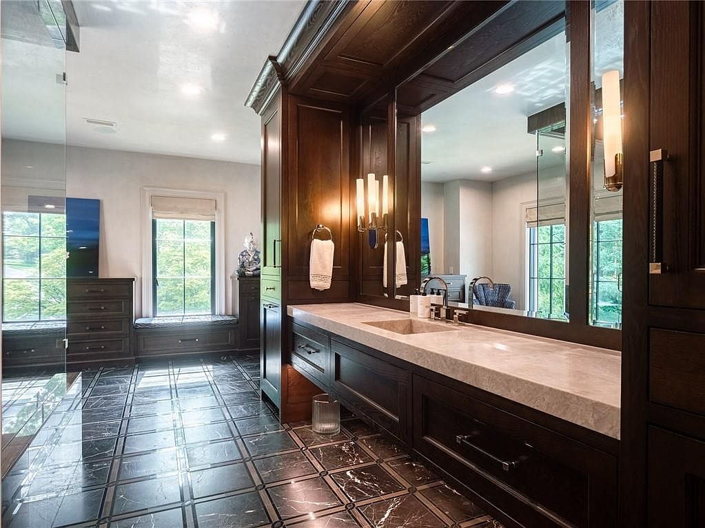 This luxurious primary bathroom features dark wood cabinetry with a light-toned stone countertop and a large, integrated mirror. The floor is covered in a sophisticated dark patterned tile, and a windowed alcove with a built-in bench provides a bright, serene seating area. The perspective is from the doorway, looking across the vanity toward the window, highlighting the elegant contrast between the dark cabinetry and the light stone surfaces.