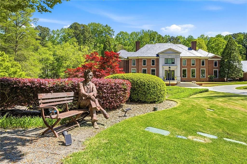 This image captures a grand, traditional brick estate set against a backdrop of lush, mature trees and a vibrant landscape. In the foreground, a bronze statue of a seated figure rests on a wooden bench, positioned near manicured hedges and a gravel path that leads toward the home's prominent stone-accented entryway. The perspective is wide and cinematic, showcasing the expansive green lawn and the architectural elegance of the residence under a clear blue sky.
