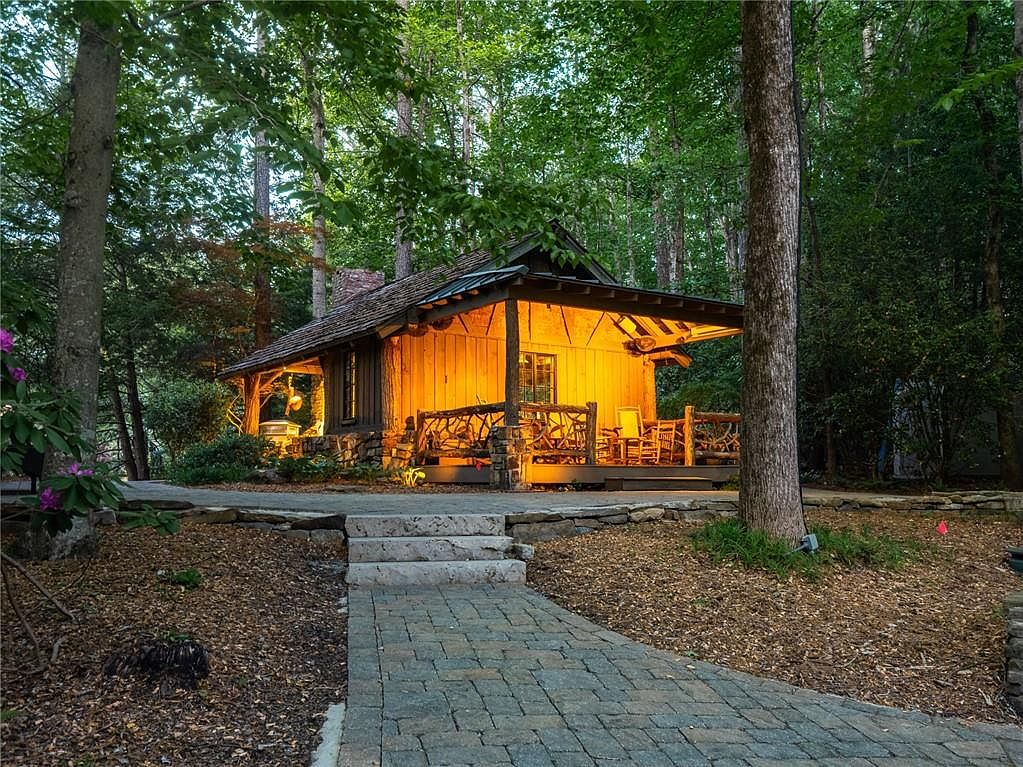 This charming rustic cabin is nestled within a lush, wooded landscape, featuring warm, inviting lighting that illuminates its wooden exterior and covered porch. A stone-paved walkway leads up to the structure, which is accented by a natural branch-style railing and a stone chimney. The scene captures a serene, cozy atmosphere, highlighting the cabin's integration with the surrounding forest.