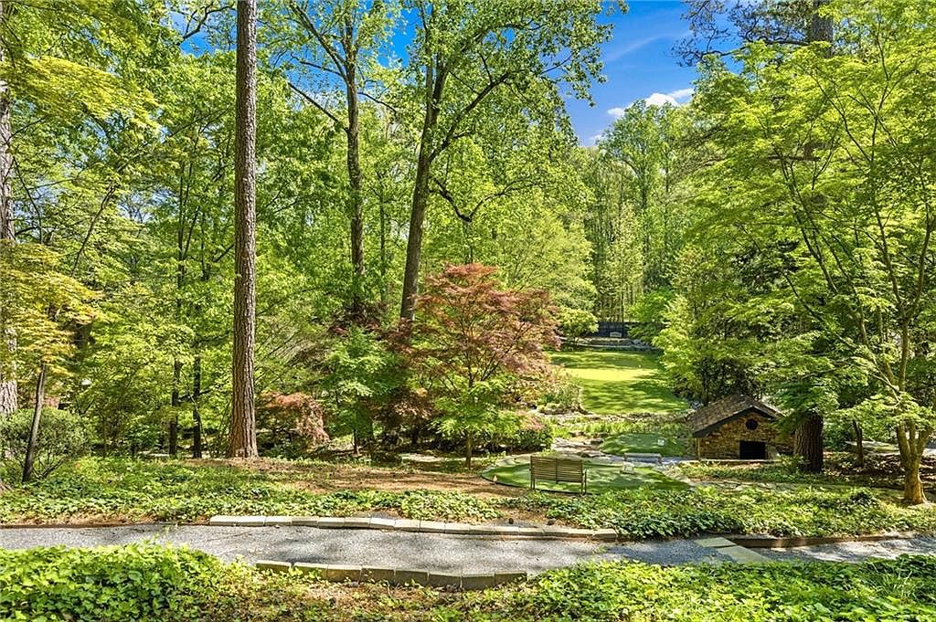 This expansive, lush backyard features a manicured putting green nestled among mature trees and vibrant foliage. A stone-clad structure sits to the right, while a wooden bench provides a peaceful seating area overlooking the green. The perspective is elevated, looking down from a gravel path onto a serene, park-like landscape that offers significant privacy and natural beauty.