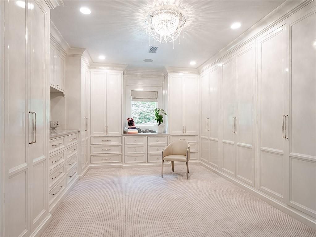 This luxurious walk-in closet features extensive white cabinetry with polished hardware, providing ample storage space. A central chair sits on the neutral-toned carpet, while a crystal chandelier hangs from the ceiling, adding a touch of elegance to the room. The space is illuminated by recessed lighting and natural light from a window centered at the back, creating a bright and organized atmosphere.