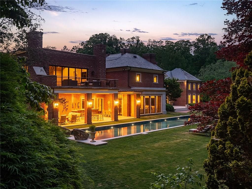 This elegant brick estate is captured at dusk, showcasing a sprawling rear facade with a covered patio and a long, rectangular swimming pool. The home features multiple levels, including a second-story balcony and large windows that glow with warm interior lighting, creating an inviting and sophisticated atmosphere. The manicured lawn and surrounding mature trees frame the property, emphasizing its private and luxurious setting.