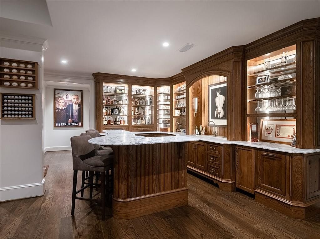 This sophisticated home bar area features rich wood cabinetry with integrated lighting and a curved island topped with a light-colored stone surface. The space is accented by dark upholstered bar stools, a collection of baseball memorabilia on the wall, and a framed political poster. The warm lighting and polished wood flooring create an inviting atmosphere suitable for entertaining.