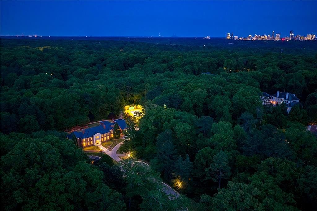 This stunning aerial night shot captures a grand estate nestled within a dense, lush forest canopy. The property features a large, illuminated residence with a winding driveway, a brightly lit tennis court, and a distant, glowing city skyline on the horizon. The perspective provides a cinematic sense of seclusion and luxury, highlighting the expansive grounds and the dramatic contrast between the dark woods and the warm, inviting lights of the home.