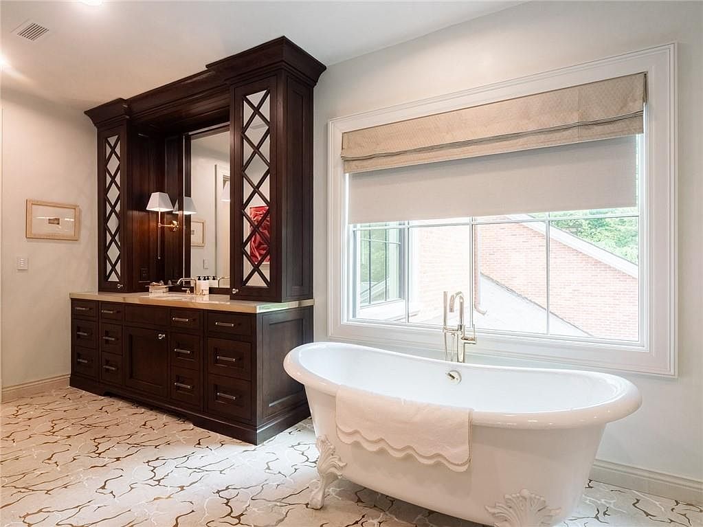 This elegant primary bathroom features a freestanding clawfoot tub positioned beneath a large window with a neutral Roman shade. To the left, a dark wood vanity cabinet is integrated into a custom built-in unit with mirrored lattice detailing and integrated lighting. The space is finished with patterned tile flooring, creating a sophisticated and tranquil atmosphere.