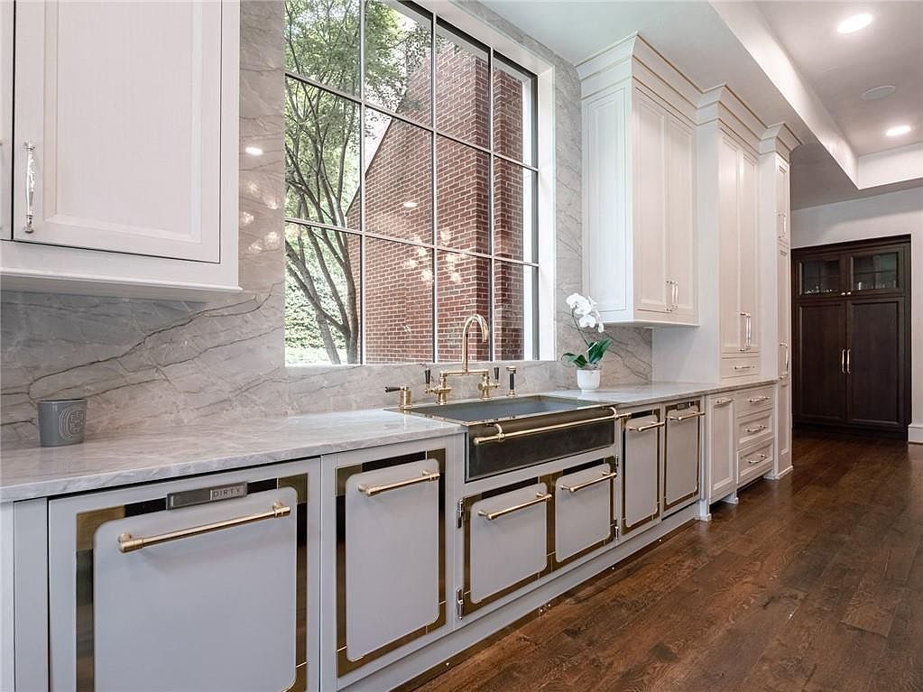 This elegant kitchen features custom light-colored cabinetry accented with polished gold-toned hardware and trim. A large, multi-pane window serves as the focal point above a farmhouse-style sink, offering a view of the exterior brickwork and trees. The space is finished with stone countertops and dark wood flooring, creating a sophisticated and high-end aesthetic.