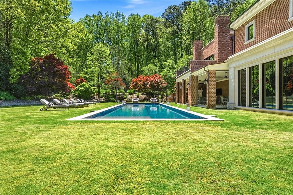 This expansive backyard features a rectangular swimming pool as the central focal point, set against a lush, tree-lined backdrop. To the right, a brick-clad home includes a covered patio area with large windows and glass doors that open to the lawn. A row of lounge chairs is neatly arranged on the grass, creating a serene and inviting outdoor living space.