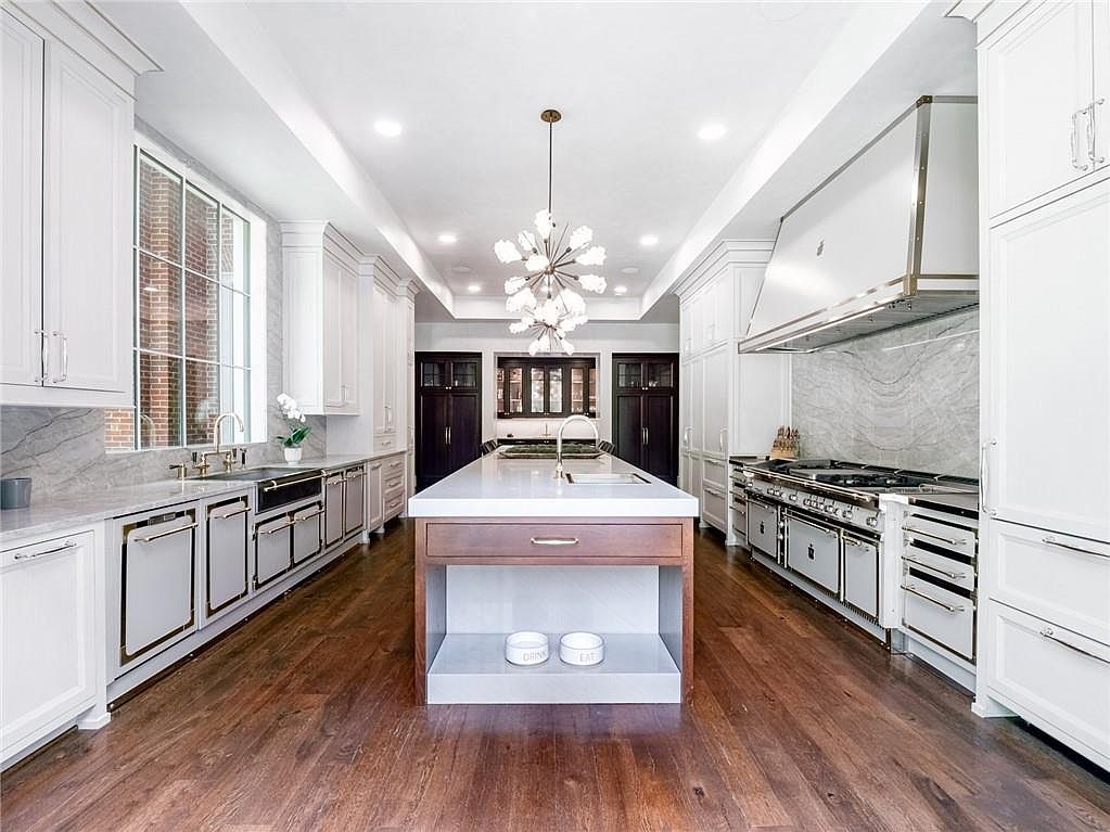 This expansive, symmetrical kitchen features white cabinetry, a large central island with a wood-toned base, and high-end stainless steel appliances. A striking modern chandelier hangs above the island, while stone countertops and backsplashes provide a sophisticated, clean aesthetic. The room is anchored by dark wood flooring and framed by dark-toned cabinetry at the far end, creating a balanced and luxurious culinary space.