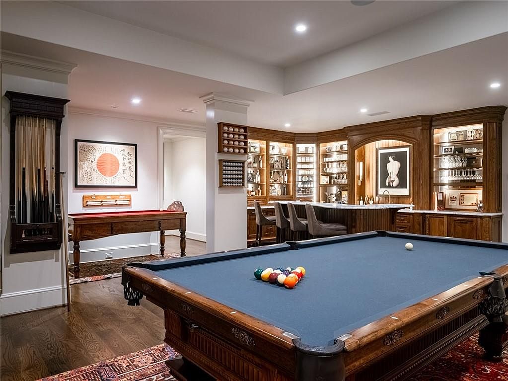 This sophisticated basement recreation room features a large pool table as the central focal point, set against a backdrop of a custom wood-paneled bar area with integrated shelving and cabinetry. The space is elegantly appointed with a wall-mounted cue rack, a secondary game table, and recessed lighting that creates a warm, inviting atmosphere. The combination of rich wood tones and classic decor gives the room a refined, club-like aesthetic perfect for entertaining.