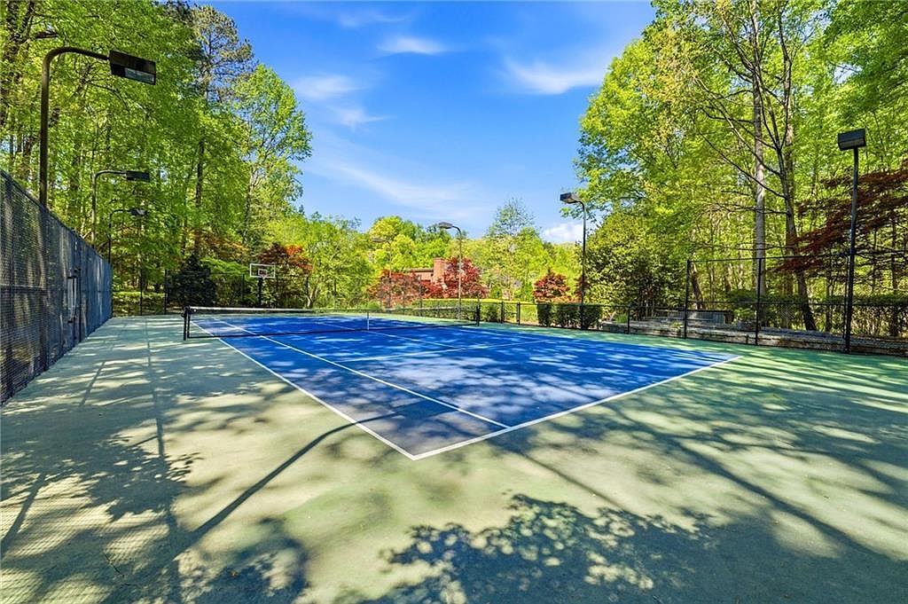 This image captures a private outdoor tennis court set against a backdrop of lush, mature trees under a bright blue sky. The court features a vibrant blue playing surface with white boundary lines and a central net, complemented by a surrounding green perimeter. Tall light poles are positioned around the court, and a basketball hoop is visible in the distance, suggesting a versatile recreational space.