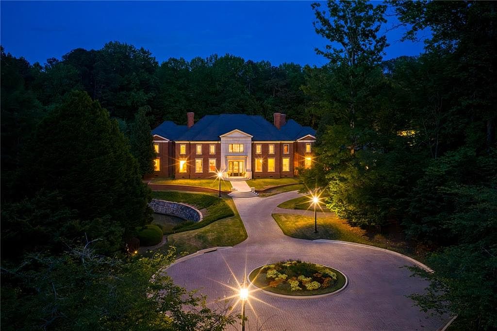 This elevated, cinematic perspective captures a grand, multi-story brick estate at dusk, illuminated by warm interior lighting and exterior landscape lamps. The property features a symmetrical facade with a prominent central stone entryway, a circular driveway with a landscaped island, and a lush, wooded backdrop. The composition emphasizes the home's stately presence and manicured grounds, creating an inviting and luxurious atmosphere.
