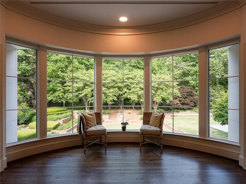 This elegant living space features a curved wall of large windows that provide a panoramic view of a lush, landscaped garden. Two matching armchairs with neutral-toned cushions are positioned symmetrically, creating a serene seating area centered around the natural scenery. The room is finished with dark wood flooring and a recessed ceiling detail, offering a sophisticated and tranquil atmosphere.