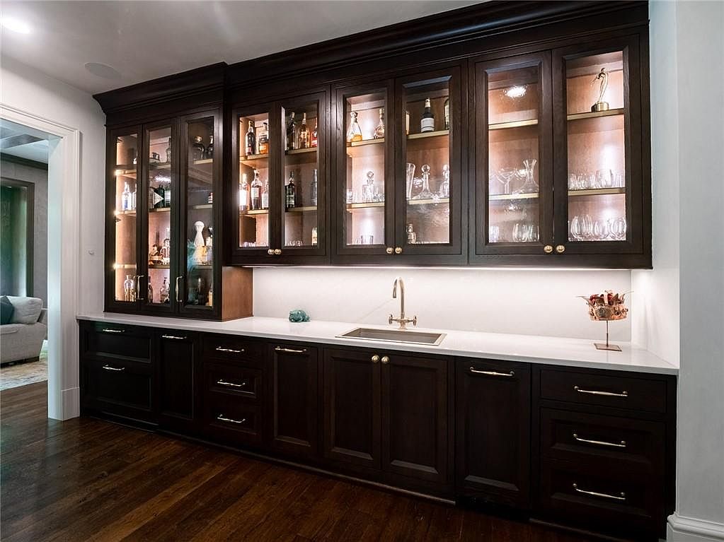This elegant wet bar area features dark wood cabinetry with glass-front upper cabinets that are illuminated from within, showcasing glassware and bottles. A sleek, light-colored countertop provides a clean workspace around a central undermount sink with a gold-toned faucet. The space is finished with dark wood flooring and gold hardware, creating a sophisticated and functional entertaining area.