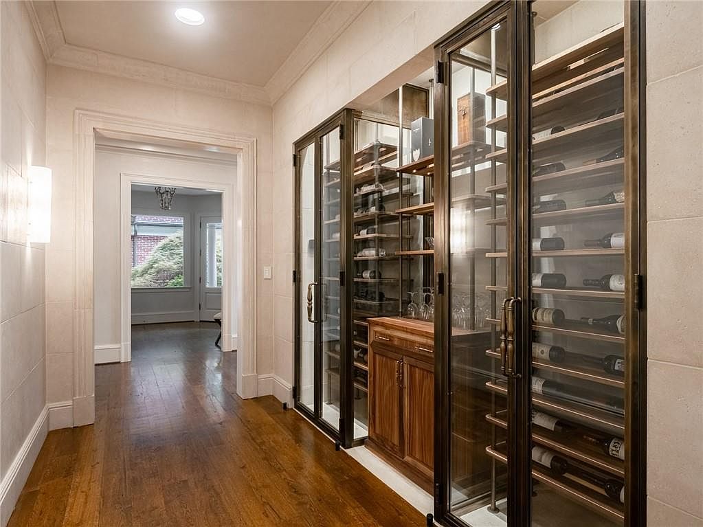 This sophisticated hallway features a custom-built, glass-enclosed wine cellar with metal racking and integrated wooden cabinetry. The space is finished with light-toned stone walls and rich hardwood flooring, creating a refined and elegant atmosphere. A doorway at the end of the hall leads to an adjacent room, providing a sense of depth and connectivity within the home.