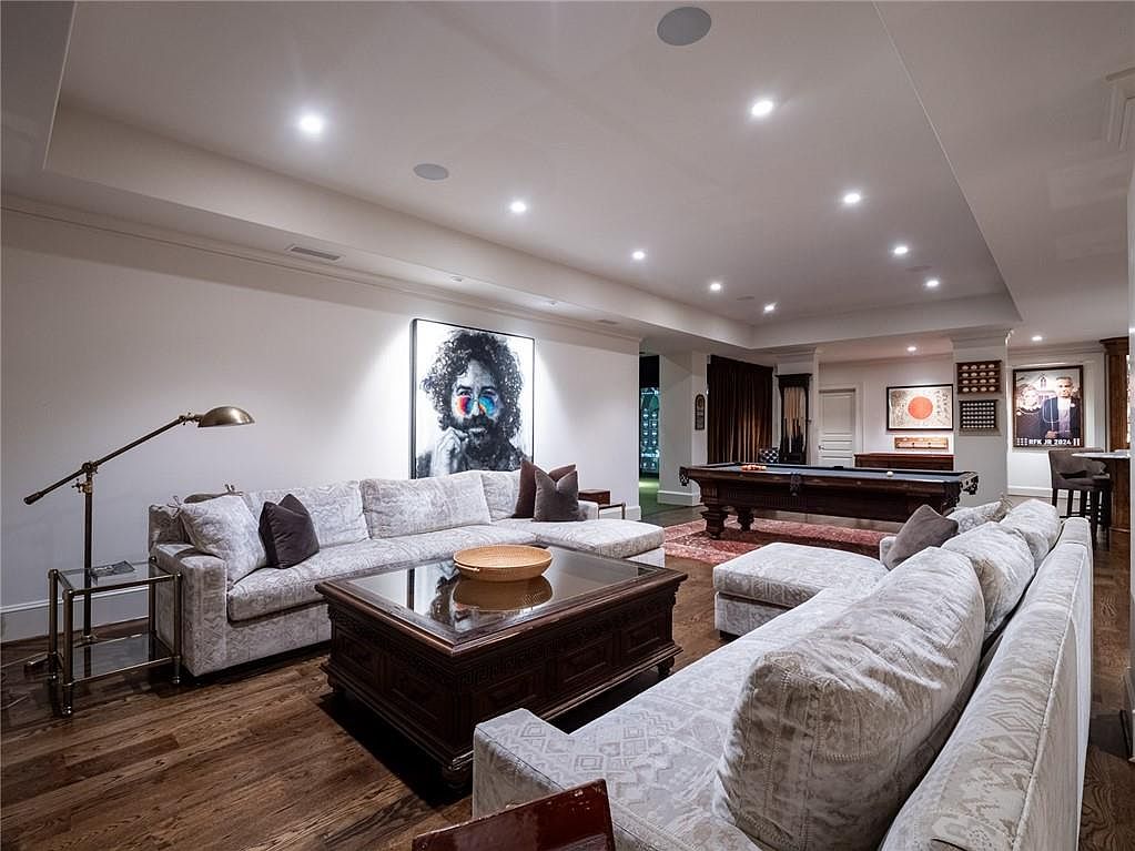 This spacious, finished basement features a large sectional sofa arranged around a dark wood coffee table, creating a comfortable lounge area. A prominent portrait of a musician hangs on the wall, while a pool table sits in the background, adding to the recreational appeal of the room. The space is illuminated by recessed lighting and includes dark wood flooring, providing a warm and inviting atmosphere for entertainment.