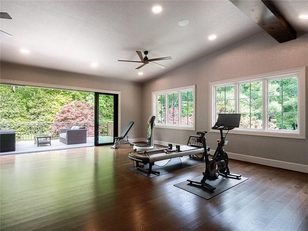 This spacious home gym features rich hardwood flooring and a neutral color palette, creating a clean and inviting workout environment. The room is equipped with a stationary bike, a Pilates reformer, and a weight bench, all positioned to take advantage of the natural light streaming through the large windows and the expansive sliding glass doors that open onto a deck. A ceiling fan provides air circulation, while the open layout offers plenty of space for various fitness activities.