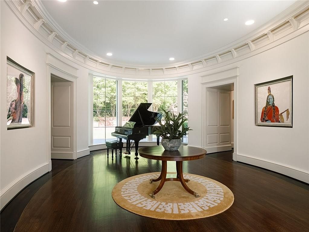 This elegant, circular-shaped room features dark hardwood flooring and a grand piano positioned in front of a curved wall of large windows. A round wooden table sits atop a patterned circular rug in the center of the space, flanked by two framed artworks on the white walls. The room is defined by its sophisticated architectural curves, recessed ceiling lighting, and a formal, gallery-like atmosphere.