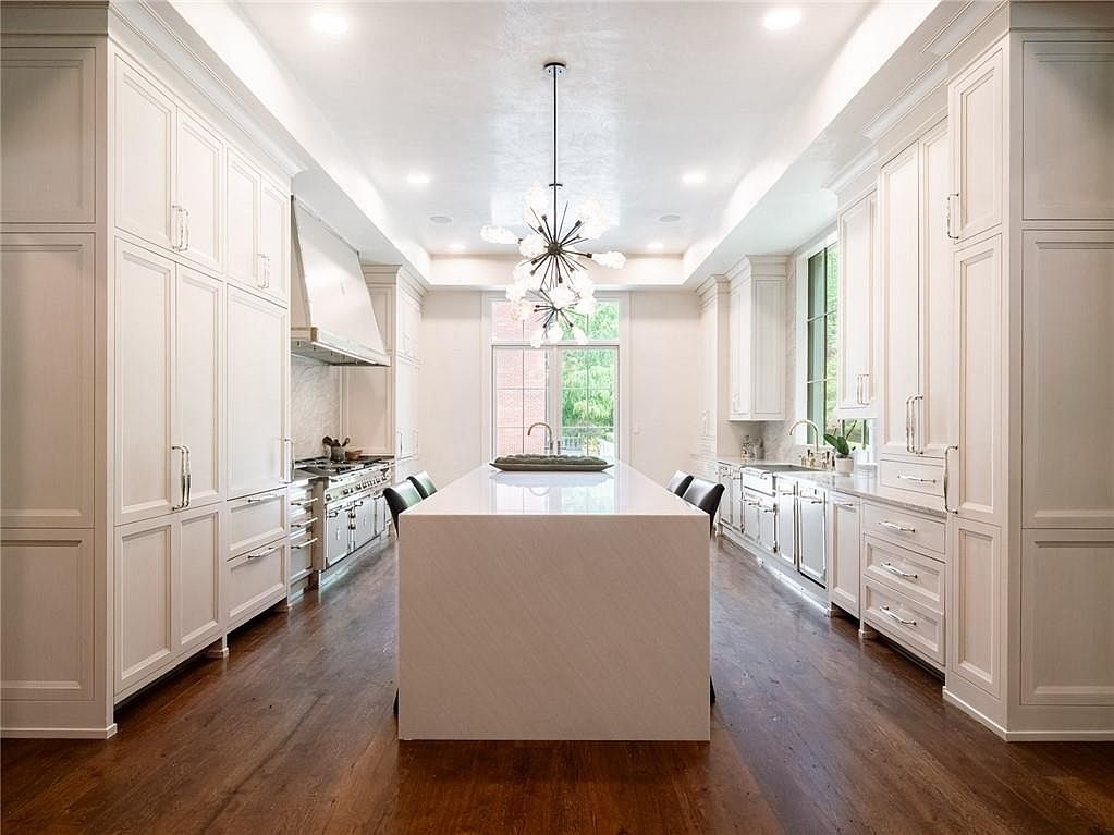 This spacious, high-end kitchen features a symmetrical layout with white cabinetry, a large central island with a stone countertop, and dark wood flooring. A striking modern chandelier hangs above the island, while a professional-grade range with a custom hood serves as a focal point on the left. The perspective is centered, looking through the kitchen toward a large glass door that allows natural light to fill the room.