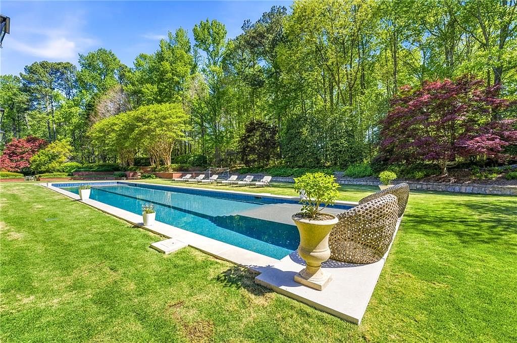This expansive backyard features a long, rectangular lap pool with a partial cover, set against a backdrop of lush, mature trees. A stone-paved pool deck surrounds the water, accented by a decorative urn planter and woven outdoor seating. The well-manicured lawn and vibrant landscaping create a serene, private retreat perfect for outdoor relaxation.