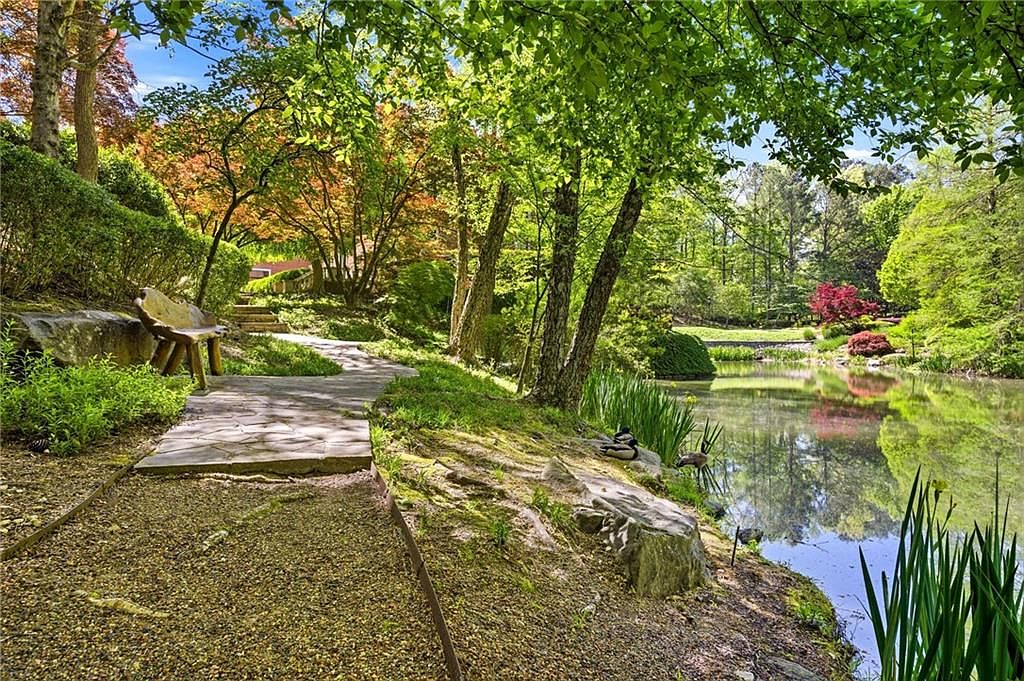This serene garden scene features a winding stone pathway leading past a rustic wooden bench nestled under mature trees. A tranquil pond reflects the lush greenery and vibrant foliage, creating a peaceful and picturesque outdoor retreat. The perspective is a wide-angle view from the path, emphasizing the natural beauty and landscaped grounds of the property.