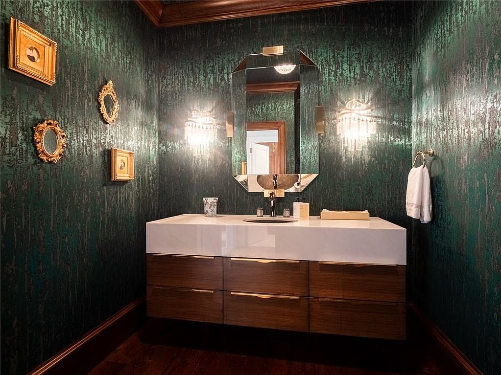 This sophisticated powder room features dramatic, textured dark green wallpaper that creates a moody and luxurious atmosphere. A floating vanity with a white countertop and horizontal wood-toned drawers is centered beneath a geometric, multi-faceted mirror flanked by elegant crystal wall sconces. The space is accented with a collection of small, ornate gold-framed art pieces on the wall, contributing to a refined and curated aesthetic.