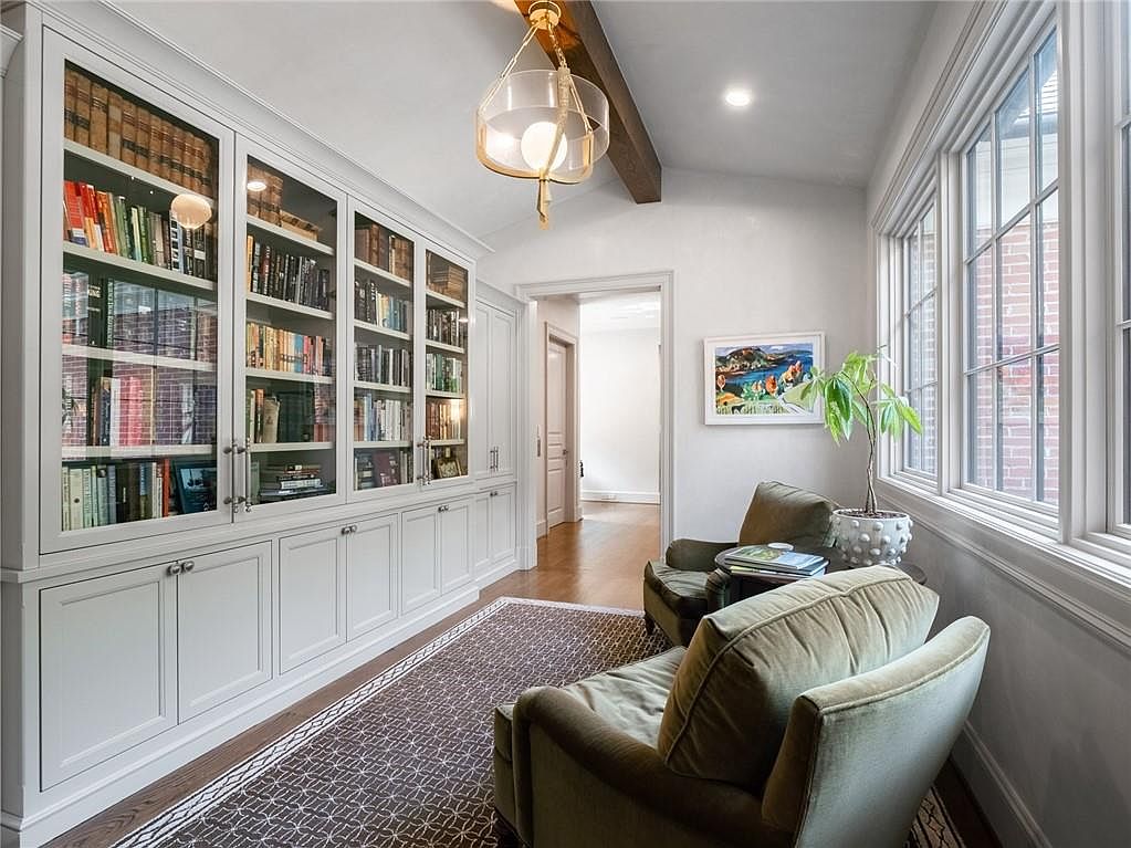 This cozy study features a wall of built-in white cabinetry with glass-fronted shelving, providing ample space for books and decor. Two comfortable olive-green armchairs are positioned near large windows that allow for natural light, while a patterned area rug adds texture to the hardwood flooring. The space is accented by a decorative pendant light and a wooden ceiling beam, creating a warm and inviting atmosphere.