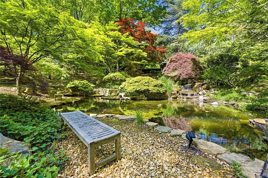 This serene garden scene features a tranquil koi pond with a small, tiered waterfall cascading over natural stone. A weathered wooden bench sits on a gravel path in the foreground, providing a peaceful vantage point to enjoy the lush, mature landscaping and vibrant Japanese maples. The perspective is a wide-angle view from the edge of the pond, capturing the harmonious blend of water, stone, and greenery.