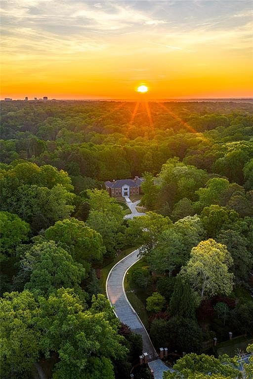 This stunning aerial perspective captures a grand estate nestled within a lush, dense forest at sunset. A long, winding paved driveway leads through the mature trees to the front of the brick residence, which is bathed in the warm, golden glow of the setting sun. The composition emphasizes the property's secluded, park-like setting and expansive grounds.