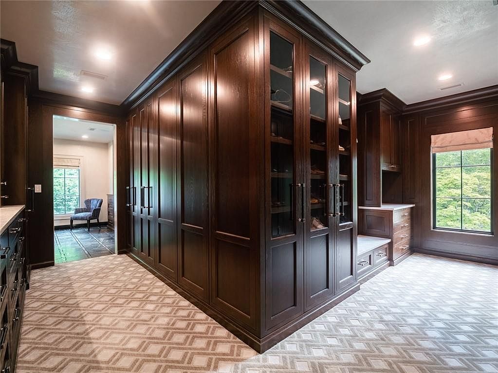 This spacious walk-in closet features extensive dark wood cabinetry with a mix of solid doors and glass-fronted display sections. A built-in bench with drawers sits beneath a window, while a patterned carpet covers the floor, adding texture to the room. The perspective captures the depth of the custom storage solutions and the transition into an adjacent room, creating an impression of luxury and organization.