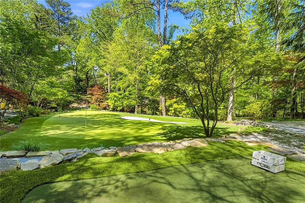 This backyard features a professionally landscaped putting green with a sand bunker, set against a backdrop of lush, mature trees. A stone retaining wall separates the putting green from the foreground, which includes a small crate for golf balls. The scene offers a serene, private atmosphere perfect for outdoor recreation.