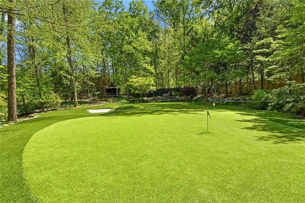 This backyard features a professionally maintained artificial turf putting green complete with multiple flag pins and a sand bunker. The green is surrounded by a lush, dense forest of mature trees, providing a private and serene atmosphere. A small wooden structure is visible in the background, adding a rustic touch to this recreational outdoor space.