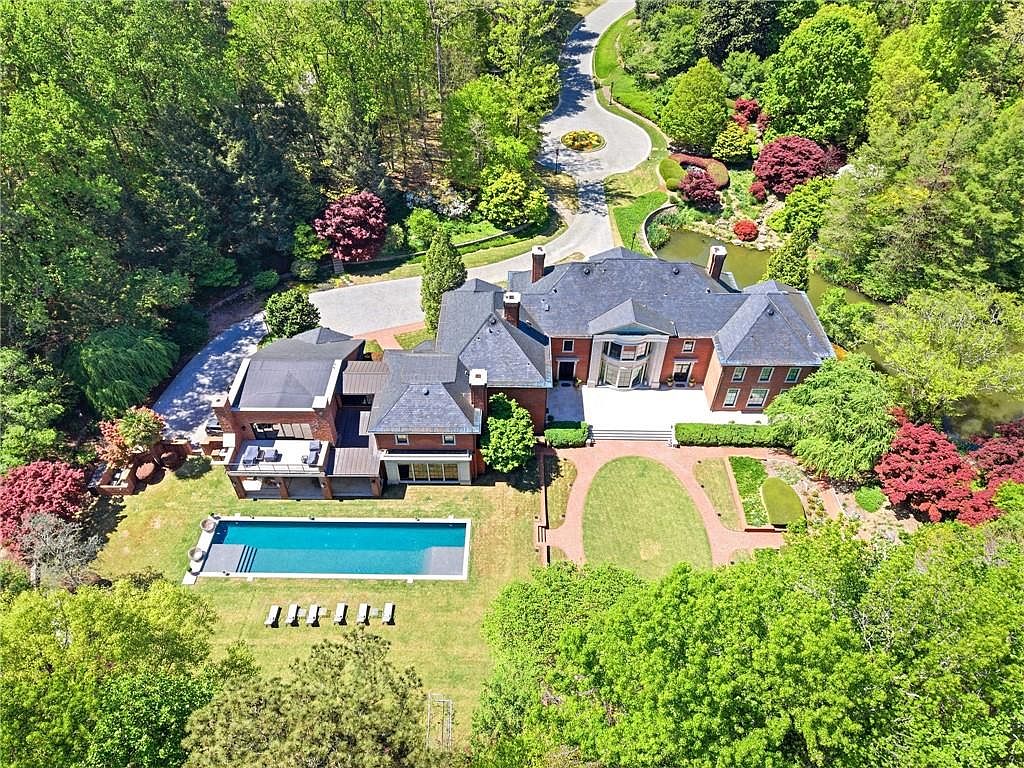 This high-angle aerial view captures a sprawling brick estate nestled within a lush, wooded landscape. The property features a rectangular swimming pool, a manicured lawn with brick-bordered pathways, and a private pond, all set against a backdrop of mature trees. The home's multi-gabled roofline and expansive rear patio area highlight its grand scale and secluded, park-like setting.