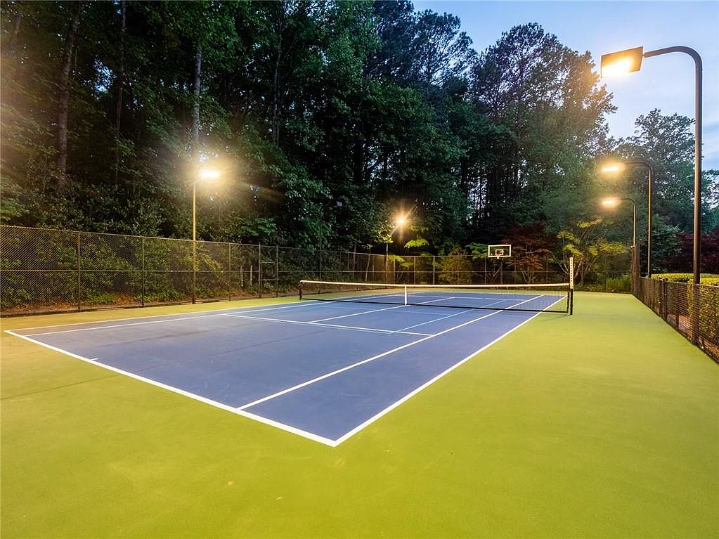 This image showcases a well-maintained private tennis court featuring a blue playing surface with contrasting green perimeter areas. The court is enclosed by high chain-link fencing and is illuminated by several tall light poles, suggesting suitability for evening play. A basketball hoop is mounted on the far fence, adding versatility to the recreational space, which is framed by a dense backdrop of mature trees.