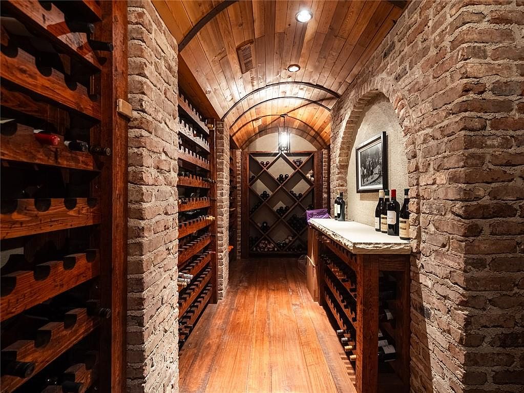 This sophisticated wine cellar features a rustic aesthetic with exposed brick walls, a barrel-vaulted wood-paneled ceiling, and warm hardwood flooring. The space is lined with extensive custom wooden wine racking, including a diamond-patterned display at the far end and a stone-topped tasting counter on the right. Recessed lighting and a hanging pendant fixture create an inviting, intimate atmosphere perfect for wine storage and appreciation.