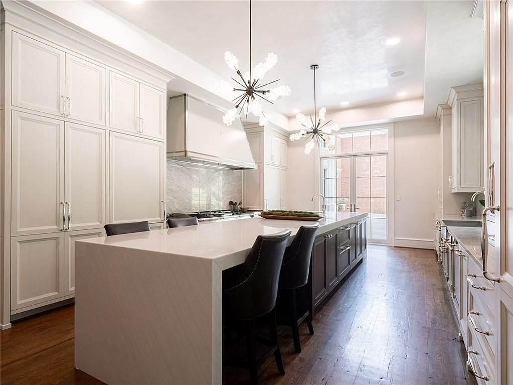 This bright, high-end kitchen features a large central island with seating, topped with a light-colored stone surface. The space is defined by white cabinetry, a professional-style range hood, and dark-toned lower cabinets, all complemented by warm wood flooring. Large glass doors at the far end provide natural light and a view of the exterior, while two modern, multi-armed chandeliers hang above the island to create a sophisticated focal point.